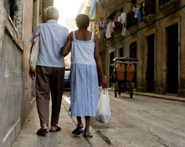 elderly man wearing a white shirt and woman wearing a blue skirt walking arm-in-arm down a narrow street
