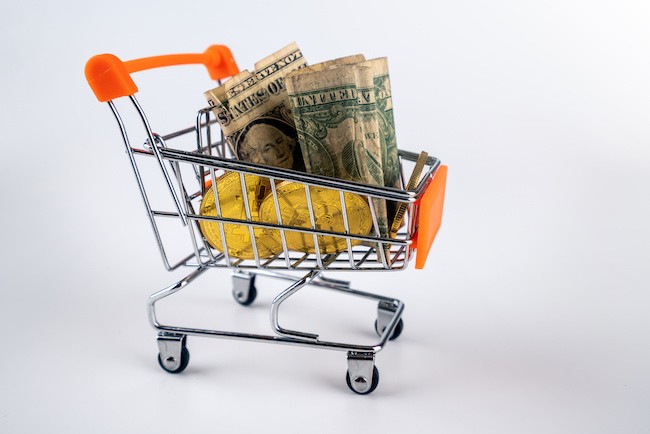 shopping cart with orange handle filled with currencies, including dollars and cryptocurrencies