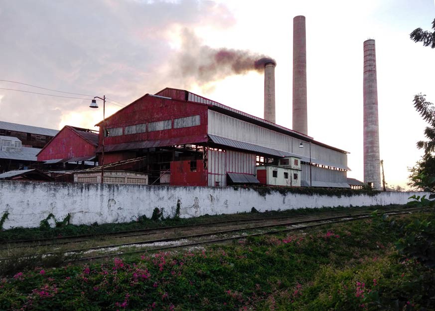 Fabrica azucarera con edificio rojo en el fondo y chimeneas echando humo y rieles de trenes en frente