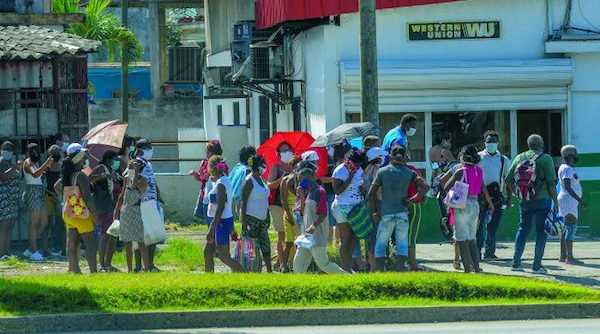 Una cola de personas con máscaras faciales, una de ellas con un paraguas rojo, en frente a un local de Western Union en Cuba.