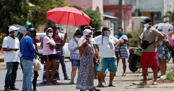 Personas portando mascarillas, esperando en cola por comida, una de ellas portando un paraguas rojo.