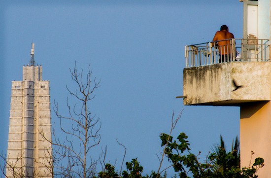 shirtless man facing back standing on a balcony perched precariously and looking at a state building