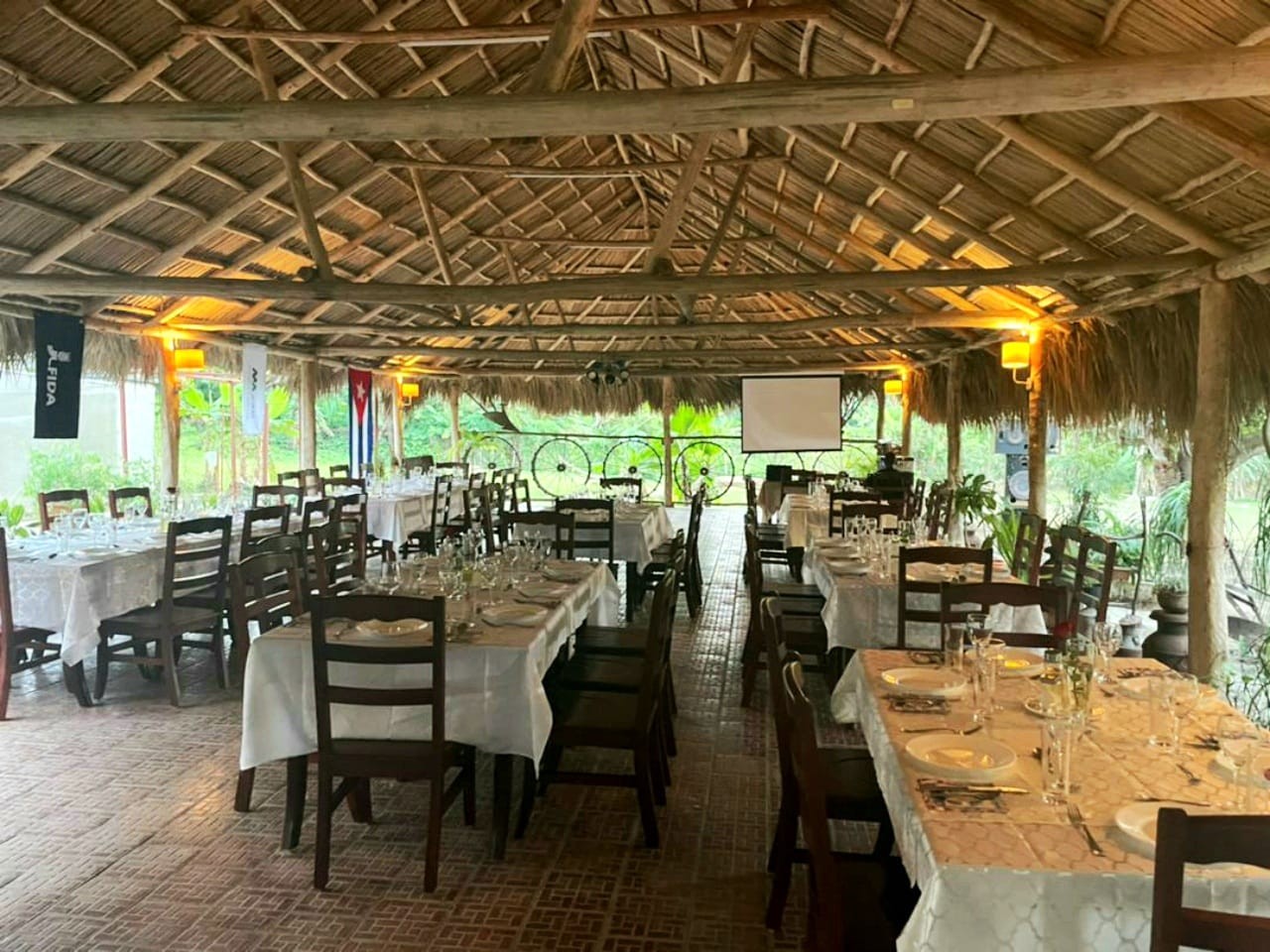 long tables with white tablecloths below a roof of natural materials and greenery in the distance.