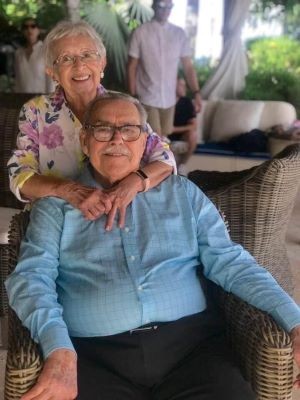 pareja sonriendo, el sentado y con camisa azul, ella detras, abrazandolo