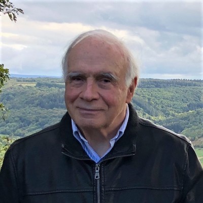 man with grey hair, smiling, wearing a dark jacket and standing before a mountainous horizon