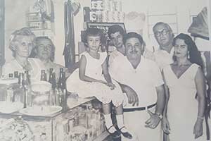 La señora mayor es la bisabuela del Sr Alvelo, MANUELA MON ALVAREZ, junto con su esposo, MANUEL CASTELAO LINARES. El más joven de la foto es su tío, ALFREDO CASTELAO MON, y las demás personas eran clientes del colmado. Grupo de personas de varias edades reunidas alrededor de un mostrador con un niña sentada encima