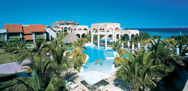 hotel complex of white buildings with pool in center, blue sky and caribbean sea in background