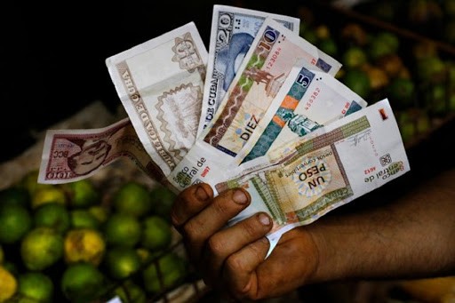 Hand with a variety of Cuban currency in front of a stand loaded with limes and fruits