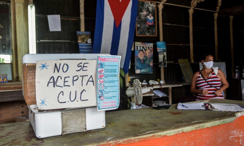 una mesa con afiche diciendo que no se aceptan CUC con una mujer sentada al lado y una bandera cubana colgada al fondo