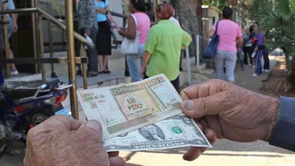 manos ancianas aguantando un peso cubano y un dólar con figuras vestidas de verde, azul claro y rosado en el fondo