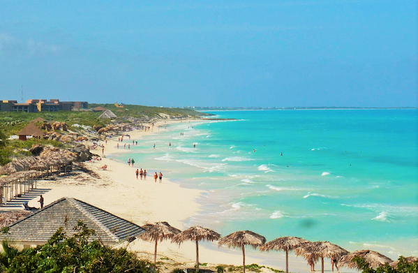 Playa de arenas blancas mar variaciones de turquesa y cielo azul con personas caminando y algunas construcciones