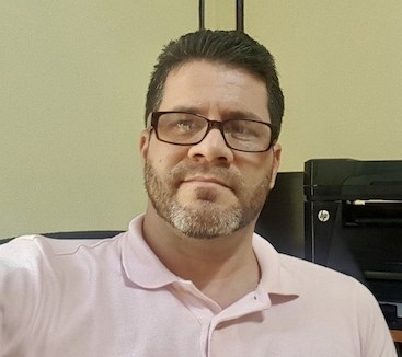 hombre con barba y lentes y pelo oscuro portando camisa rosada con impresora atrás