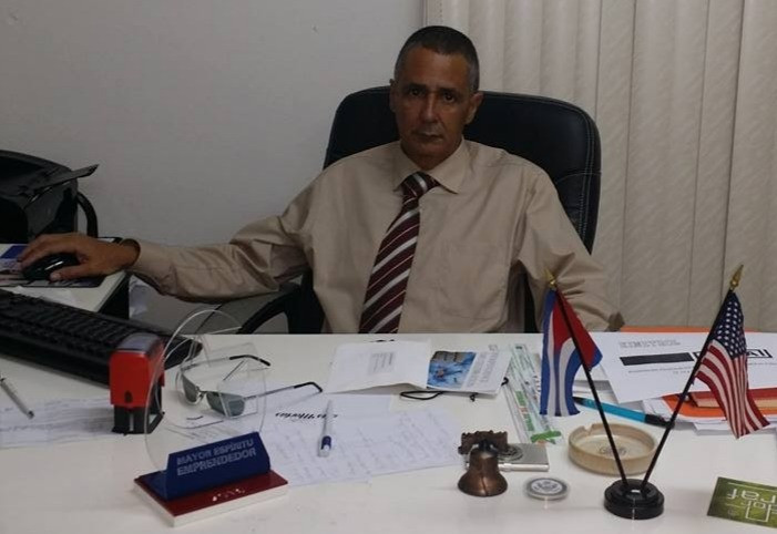 Un hombre vestido con camisa beige y corbata a rayas rojas está sentado en un escritorio de oficina. Frente a él hay documentos, una computadora y dos pequeñas banderas, una de Cuba y otra de Estados Unidos. La escena transmite el ambiente de trabajo y emprendimiento en un entorno empresarial.