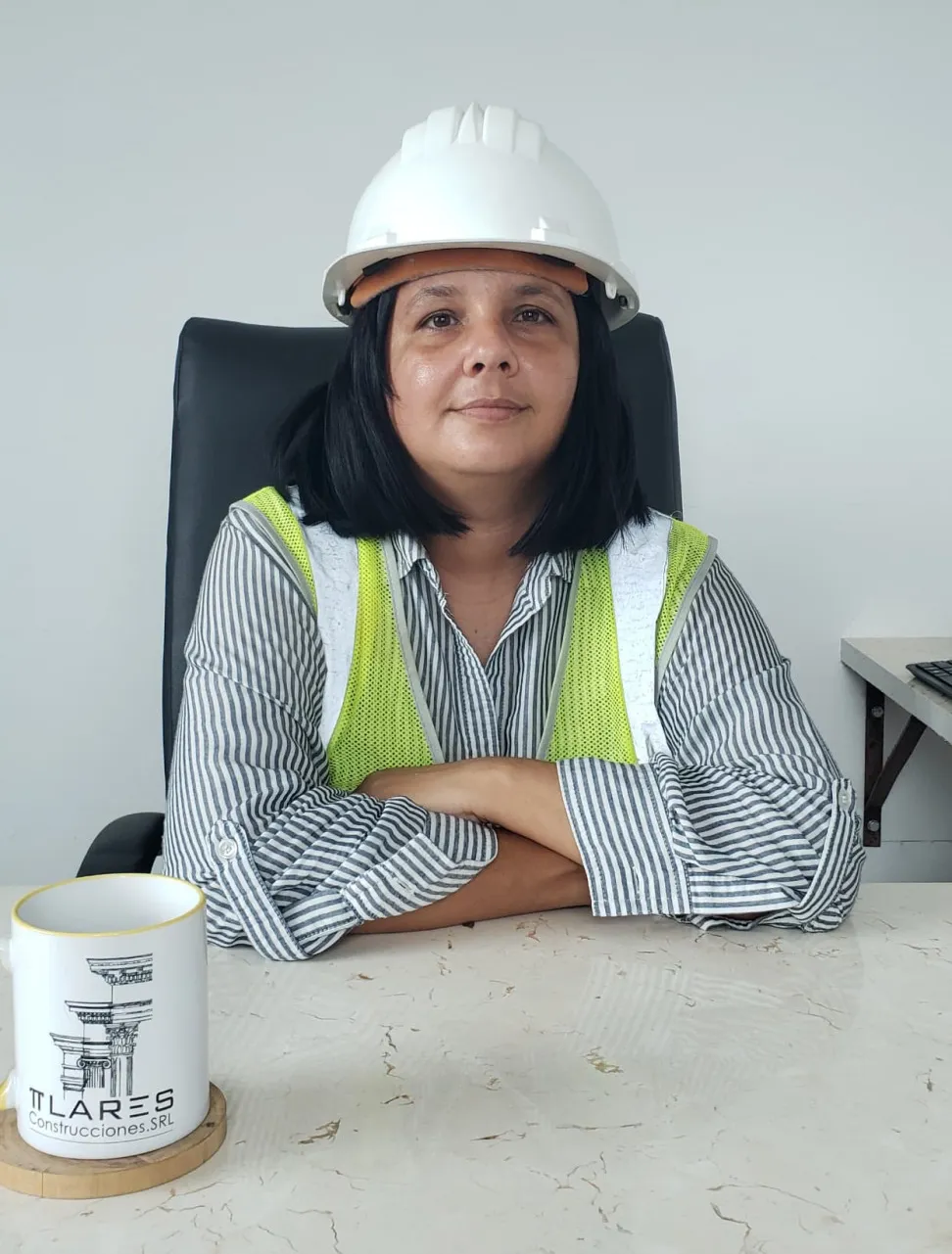 Woman wearing a construction vest and helmet, sitting with her arms crossed on a work table, inside an office.