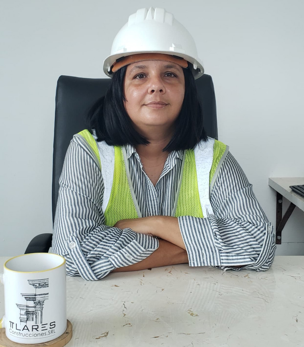 Mujer vestida con chaleco y casco de construcción, sentada con los brazos cruzados sobre una mesa de trabajo, dentro de una oficina.