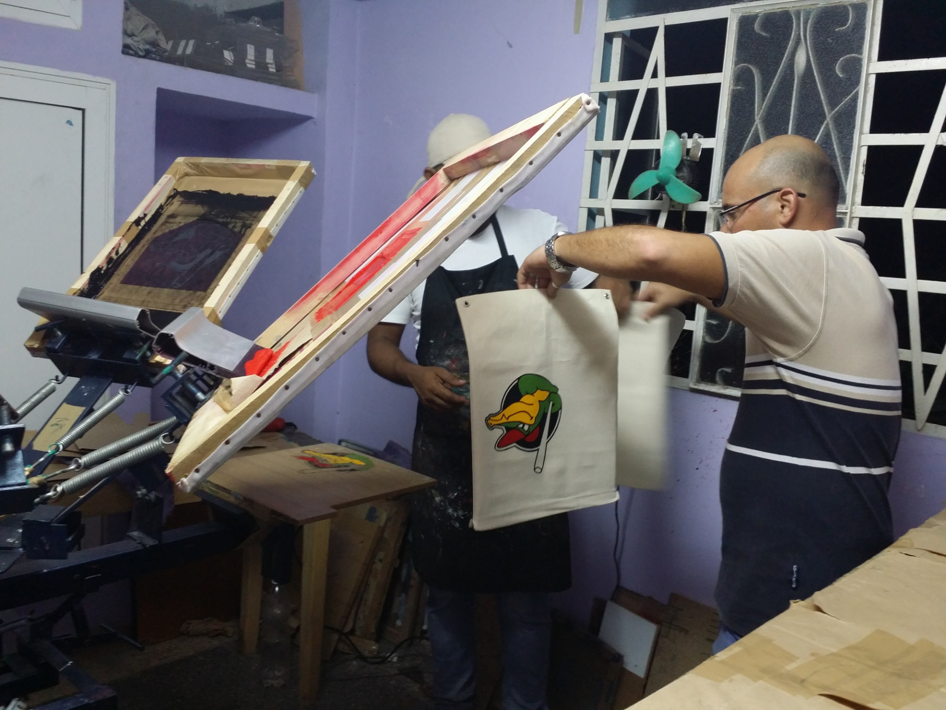 Dos hombres, en Cuba, trabajan en un rudimentario taller de serigrafía. Uno sostiene una bolsa de tela con un logotipo colorido, mientras el otro maneja una máquina de impresión con marco de madera.