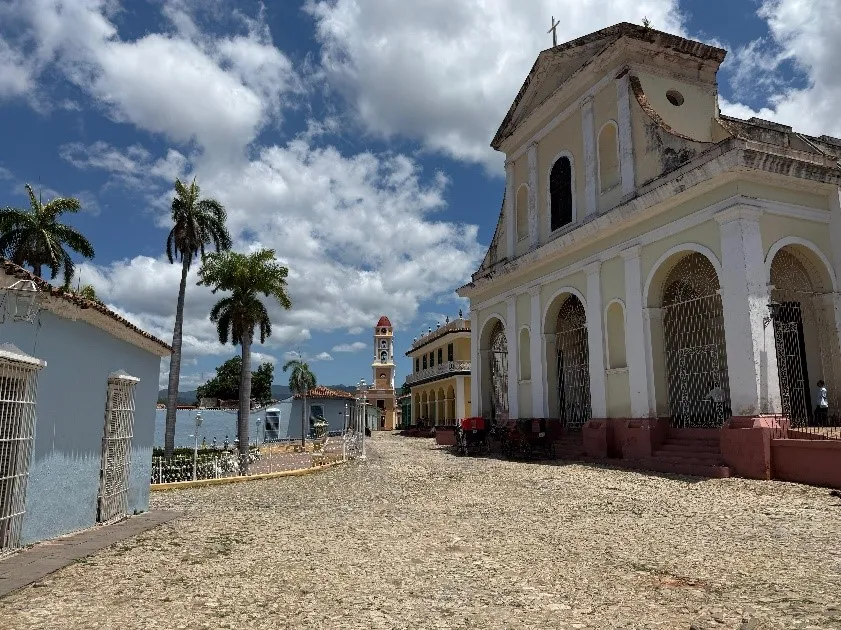 Calle empedrada y soleada de una ciudad colonial con una iglesia grande, casas coloridas, palmeras y una torre con reloj al fondo bajo un cielo azul con nubes.