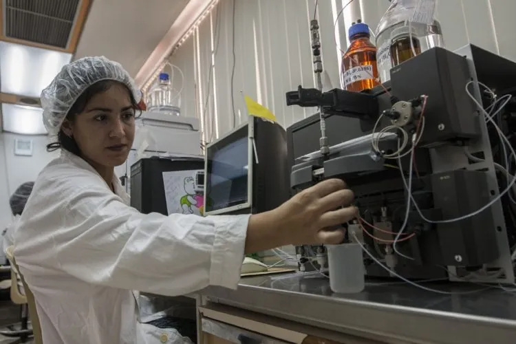 Una mujer joven vestida con bata y gorro de laboratorio manipula equipos científicos dentro de un centro de investigación. Frente a ella hay instrumentos y recipientes químicos conectados por tubos y cables. La escena transmite trabajo científico y participación femenina en áreas tecnológicas y de investigación en Cuba.