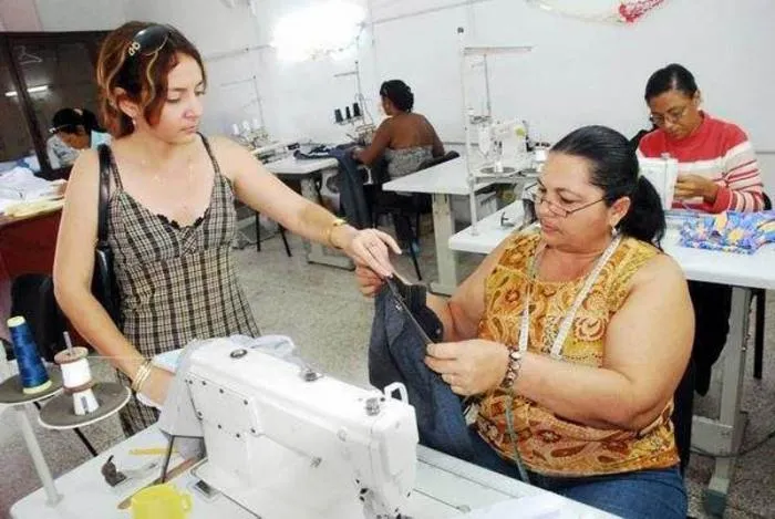 La imagen muestra un espacio de trabajo tipo taller de costura o confección. En primer plano, una mujer joven de pie interactúa con otra mujer sentada que está utilizando una máquina de coser industrial. Al fondo, se observan otras dos mujeres realizando labores de costura en sus propias estaciones de trabajo, lo cual subraya la predominancia femenina en este entorno laboral específico.