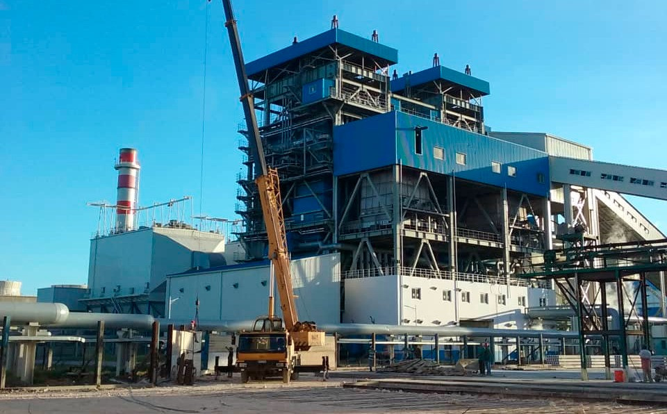 Vista exterior de una planta bioeléctrica con una estructura industrial de varios niveles en colores azul y blanco. En primer plano se observa una grúa móvil y un sistema de tuberías elevadas; al fondo, una chimenea con franjas rojas y blancas se eleva frente a un cielo despejado.