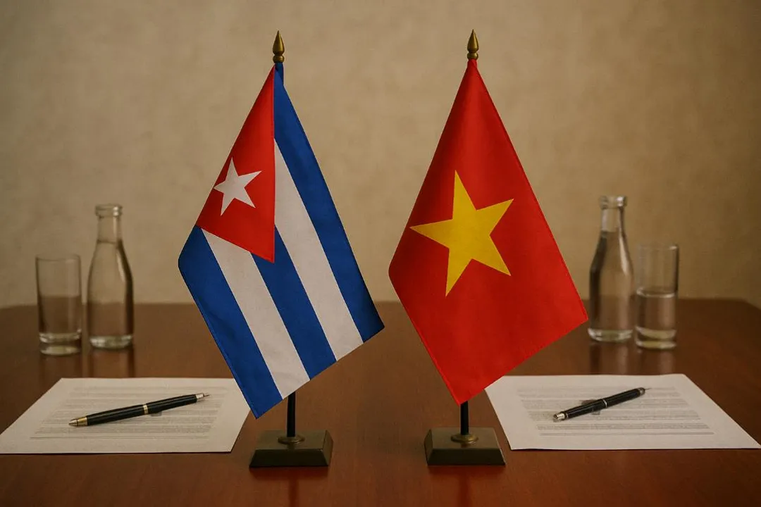En una mesa de madera pulida hay dos pequeñas banderas colocadas una al lado de la otra. A la izquierda está la bandera de Cuba, con franjas azules y blancas y un triángulo rojo con una estrella blanca. A la derecha está la bandera de Vietnam, roja con una gran estrella amarilla en el centro. Frente a cada bandera hay un documento con un bolígrafo y, más atrás, botellas y vasos de agua. El ambiente transmite formalidad y cooperación entre ambos países.