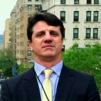 Un hombre con traje oscuro, camisa azul clara y corbata amarilla posa al aire libre frente a una avenida urbana. Al fondo se observan edificios de varios pisos, árboles y una calle con tráfico, en un entorno de ciudad.