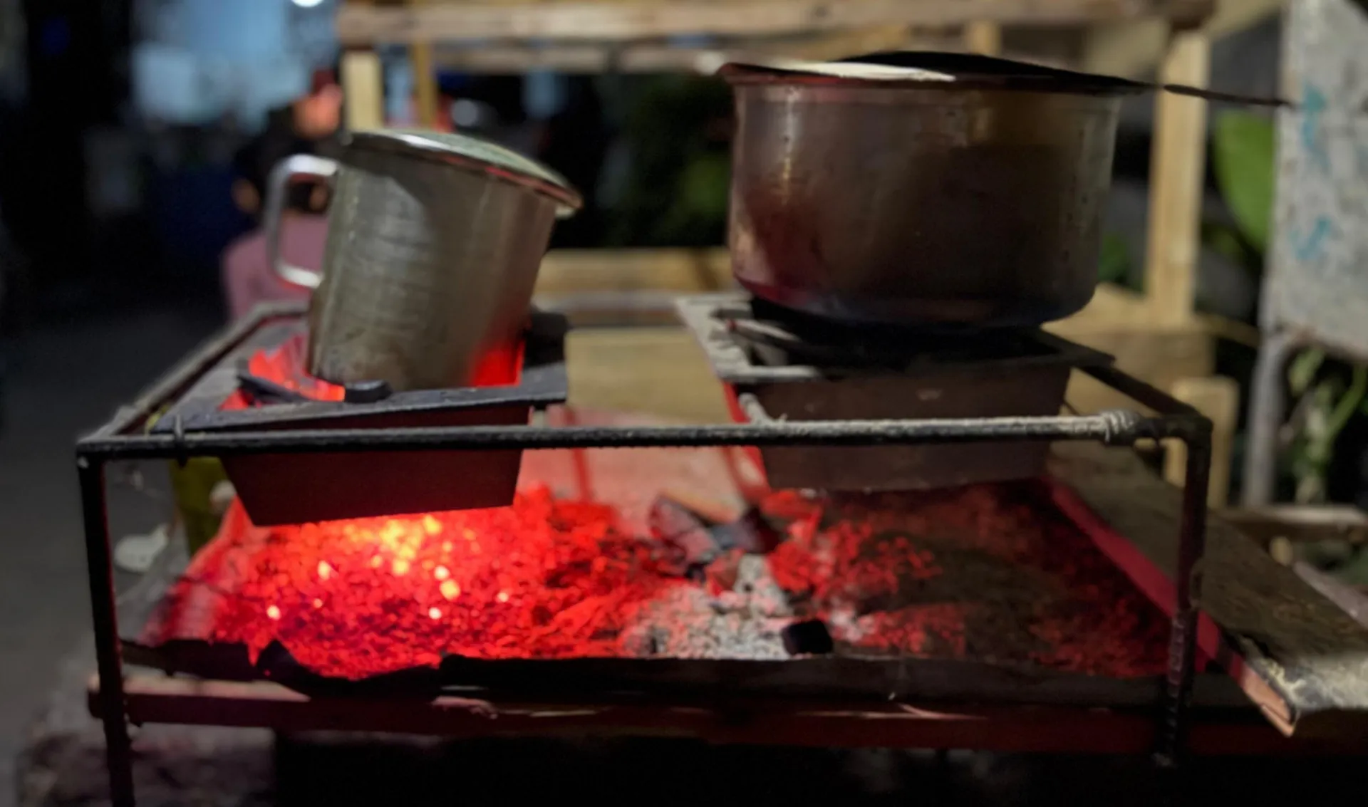 The image shows an improvised charcoal stove used for cooking, built from a rectangular metal frame. Inside, glowing red embers burn brightly, casting light across the scene. On top of the stove rest two metal containers: on the left, a tilted jug with a handle, and on the right, a large lidded pot, both supported by metal grates.