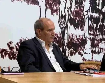 A man wearing a dark jacket and a white shirt is seated at a wooden table during a meeting or conference. He is looking intently to the side with a serious expression. On the table, there are documents, a pair of glasses, and a mobile phone. In the background, an artistic mural in shades of white, black, and brown is visible.