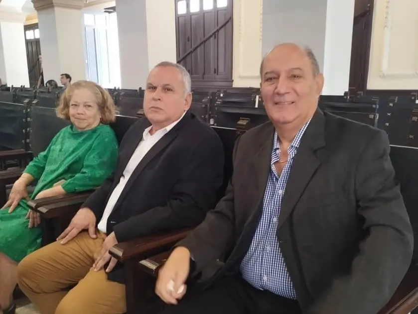 Three people are seated on benches in a spacious colonial-style hall. On the left, a woman wears a green outfit; in the center, a man in a dark jacket, white shirt, and beige trousers maintains a serious expression; on the right, another man smiles, dressed in a dark jacket and a blue-checked shirt. In the background, rows of wooden benches and large wooden doors are visible, creating an institutional atmosphere.