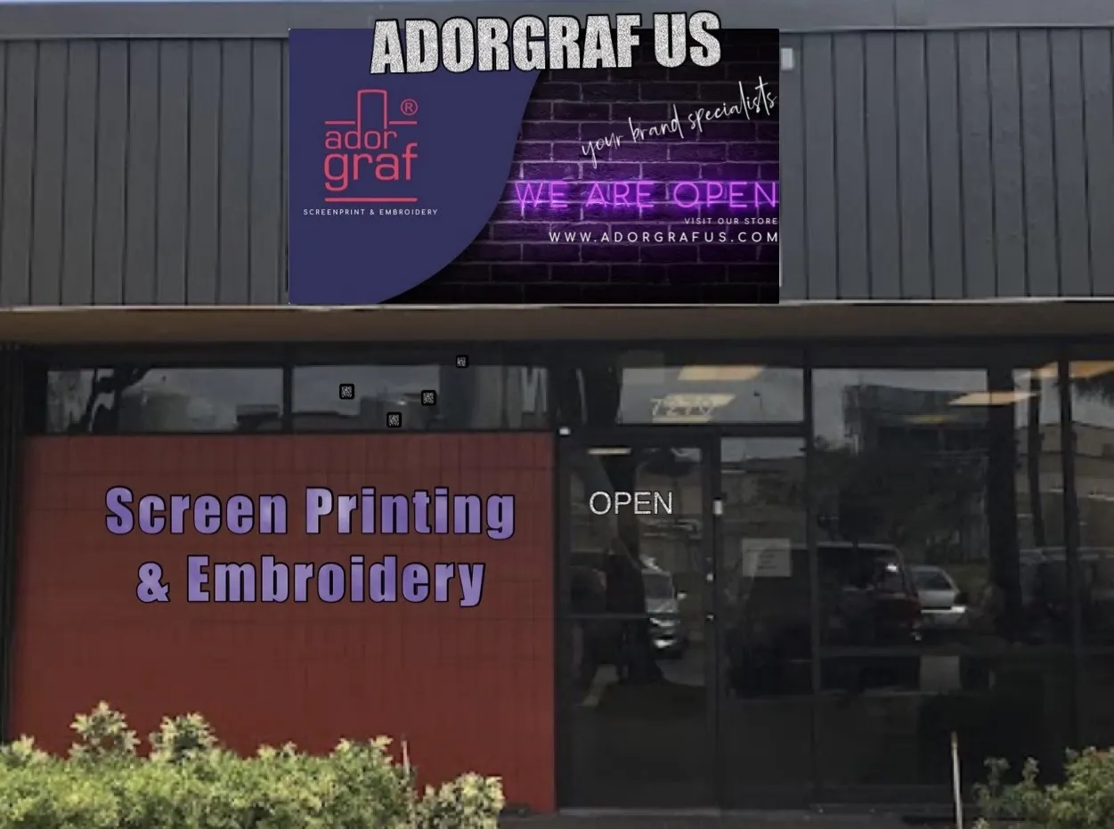 Fachada de un local comercial con un cartel superior que dice “ADORGRAF US” y muestra el logotipo de la empresa. En la pared se leen las palabras “Screen Printing & Embroidery” (impresión en serigrafía y bordado). La puerta de vidrio tiene un letrero luminoso con la palabra “OPEN” (abierto).