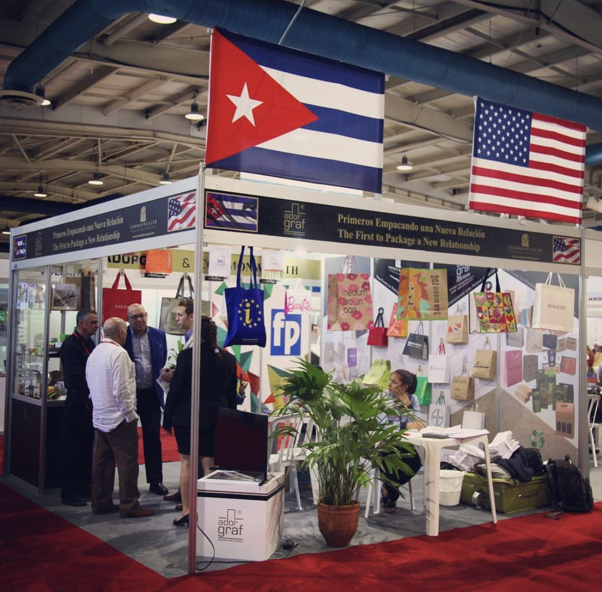 Stand de feria de la empresa Adorgraf y Commonwealth Packaging Company, decorado con las banderas de Cuba y Estados Unidos.  Se exhiben bolsas impresas y otros productos de estas empresas.