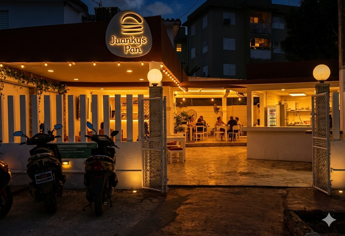 Fachada de un pequeño negocio llamado “Juanky’s Pan” iluminado cálidamente durante la noche. El local tiene un diseño abierto, con mesas donde varias personas conversan y comen. En la entrada hay dos motocicletas estacionadas y una reja blanca abierta. Al fondo, se ven edificios residenciales bajo un cielo oscuro con estrellas, creando un ambiente acogedor y tranquilo.