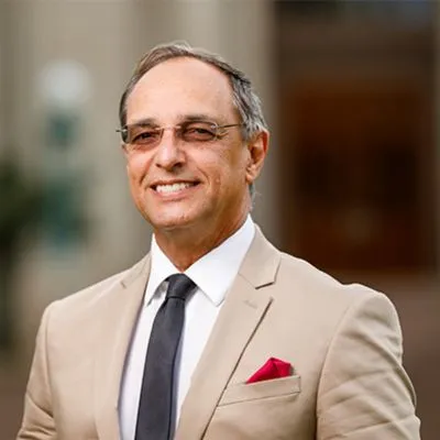 Hombre sonriente con gafas, vestido con un traje claro y corbata oscura, con un pañuelo rojo en el bolsillo.
