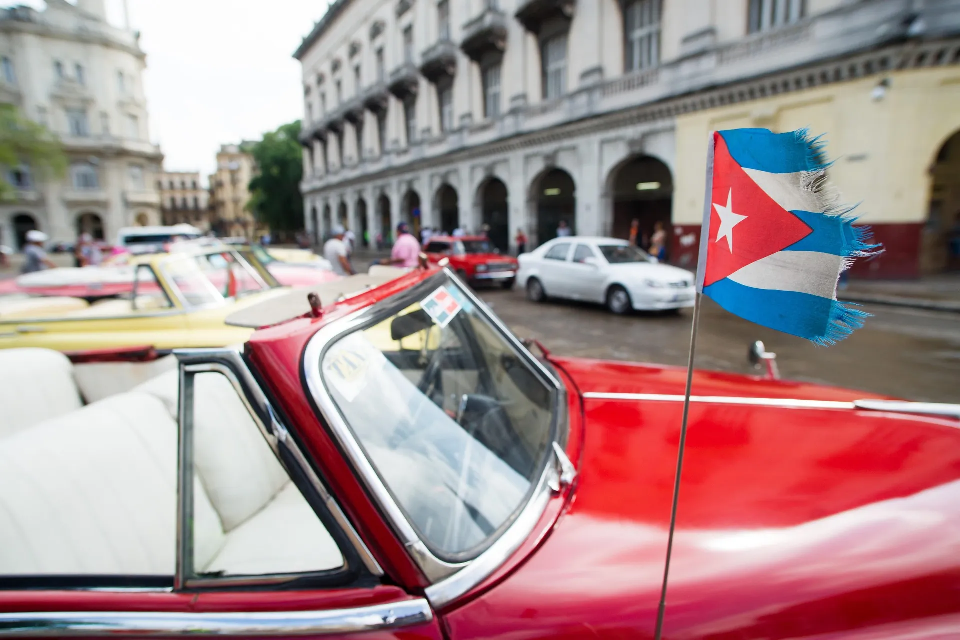 Photograph of a street in the historic center of Havana. In the foreground is a red classic American car, viewed from the side, with a white leather interior. A small Cuban flag is mounted on the car; it has blue and white stripes, a red triangle, and a white star, with worn and frayed edges. In the background are several other classic cars in different colors, along with modern vehicles and pedestrians walking along the street.