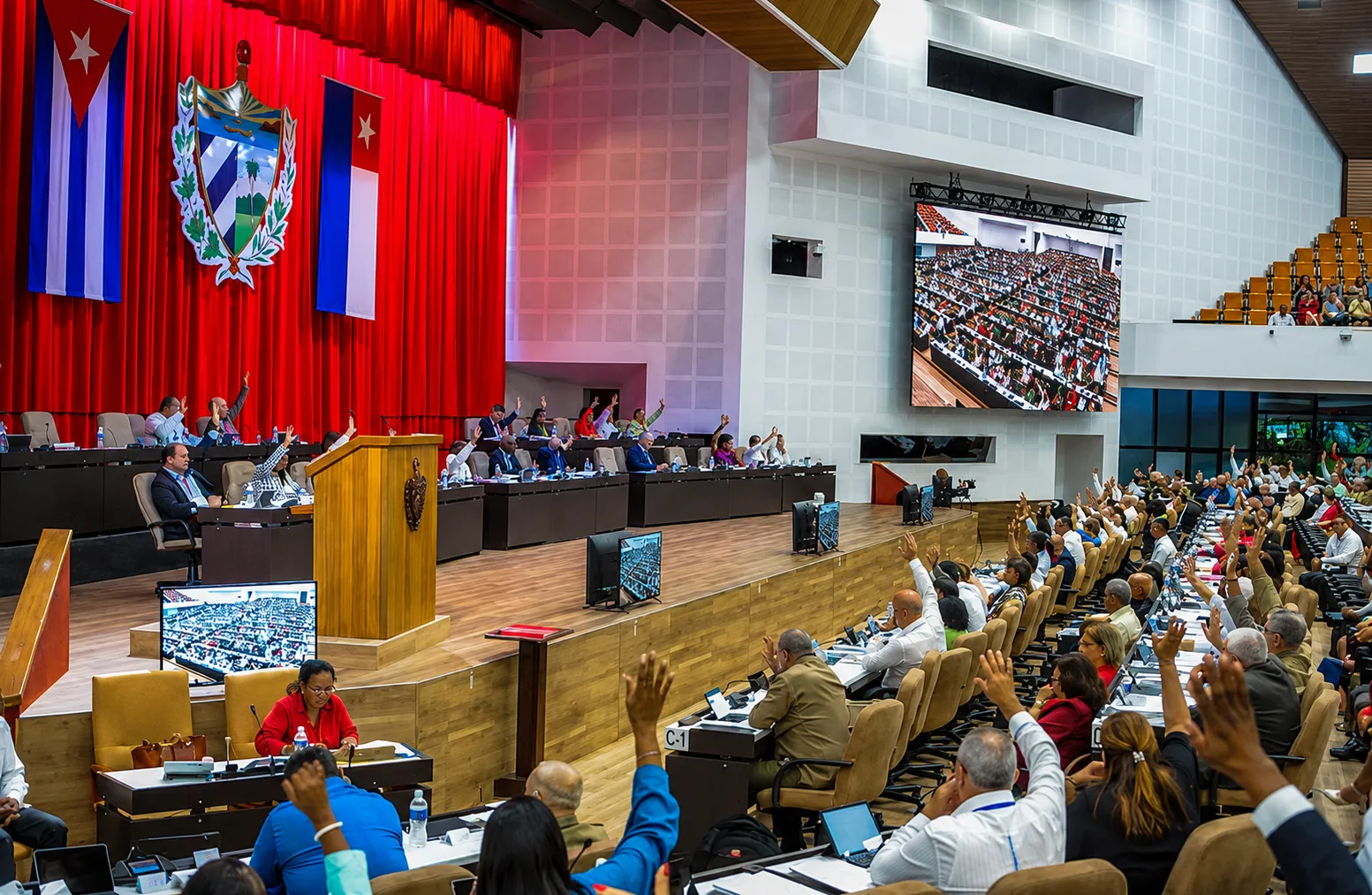 Panoramic view of the chamber of Cuba’s National Assembly of People’s Power during a parliamentary session. Numerous deputies are seated in rows of desks, with several raising their hands during a vote. In the background are the presiding table, Cuban flags, the national coat of arms, and screens displaying the chamber.