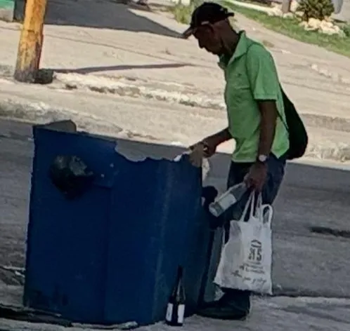 The image shows a man wearing a dark cap and light green T-shirt, leaning toward a blue trash can. He is carrying a white bag in one hand and an object in the other, while a glass bottle lies on the ground next to the container.