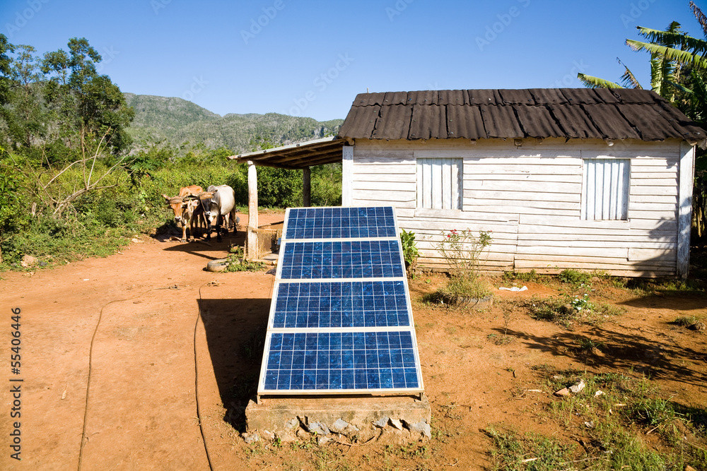 Photograph of a rural setting in Cuba. In the foreground, several tilted solar panels are installed on a concrete base on reddish soil. In the background, there is a small white-painted wooden house with a dark roof. To the left, near a shed, two oxen are tied. The landscape is surrounded by abundant vegetation and mountains under a clear sky, illustrating the use of solar energy in a rural community.