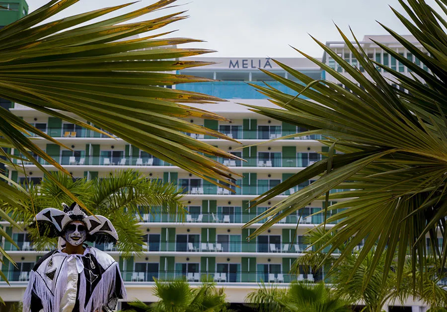 Photograph of the Meliá Internacional Varadero Hotel, a modern multi-story building with outward-facing balconies. In the foreground are tropical palm trees and a person dressed in a traditional harlequin costume. The image illustrates the high-standard hotel infrastructure built in Cuba over the past several years.