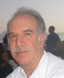 Close-up portrait of a mature man with short, greying hair, a clear forehead, brown eyes, and a well-groomed greyish mustache above his lips. He is wearing a white shirt and looking directly forward with a calm, serious expression. In the blurred background, the silhouettes of other people can be seen outdoors under soft lighting.