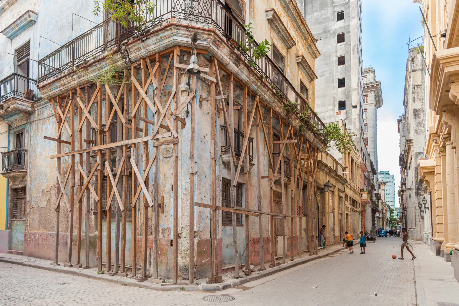 Una calle estrecha en La Habana Vieja muestra edificios antiguos y desgastados. En la esquina, una construcción de dos plantas está muy deteriorada y sostenida por una estructura de numerosos puntales de madera que cubren toda la fachada. Las paredes presentan grietas, pintura descascarada y tonos apagados de azul y ocre. En el lado derecho de la imagen, varios niños y adolescentes juegan en la calle con una pelota, mientras otros se apoyan en las paredes deterioradas.