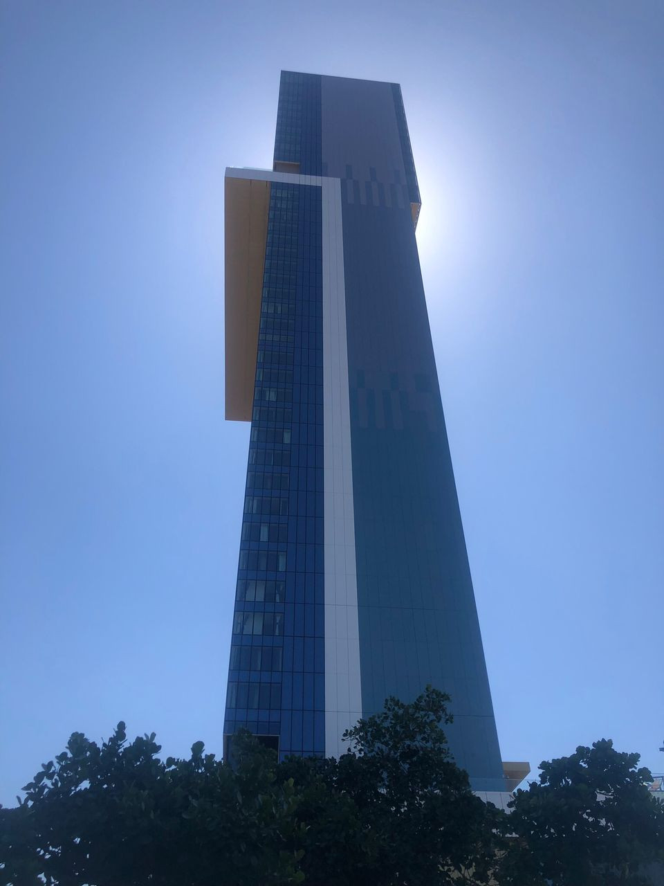 La imagen muestra un rascacielos moderno de color azul oscuro y blanco, visto desde su base hacia el cielo. La estructura es alta, de líneas rectas y paneles de vidrio reflectantes, con un diseño que incluye una sección sobresaliente en la parte superior. El cielo es completamente despejado, de un azul intenso, y el sol se encuentra justo detrás del edificio, generando un halo brillante alrededor de su cima. En la parte inferior se aprecian las siluetas de algunos árboles oscuros que enmarcan la base de la 