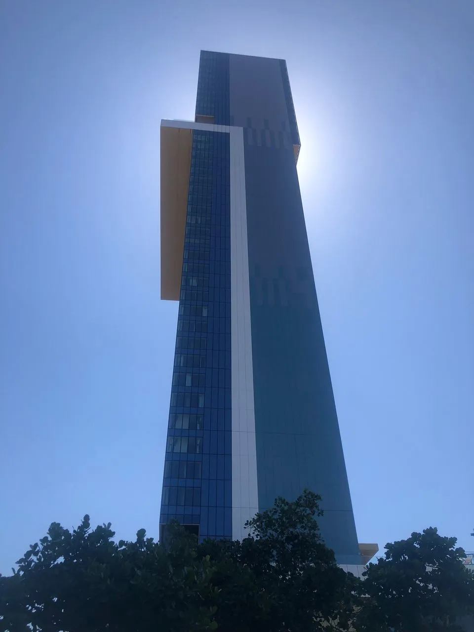 The image shows a modern skyscraper in dark blue and white, seen from its base looking up toward the sky. The structure is tall, with straight lines and reflective glass panels, featuring a protruding section at the top. The sky is completely clear, an intense blue, and the sun is positioned directly behind the building, creating a bright halo around its summit. At the bottom, the silhouettes of several dark trees frame the base of the building.