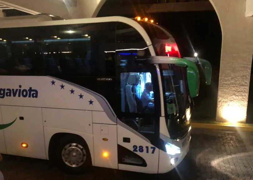 Se muestra un autobús turístico blanco de la compañía Gaviota, estacionado de noche bajo una estructura iluminada, con sus luces encendidas y el conductor visible a través del parabrisas.