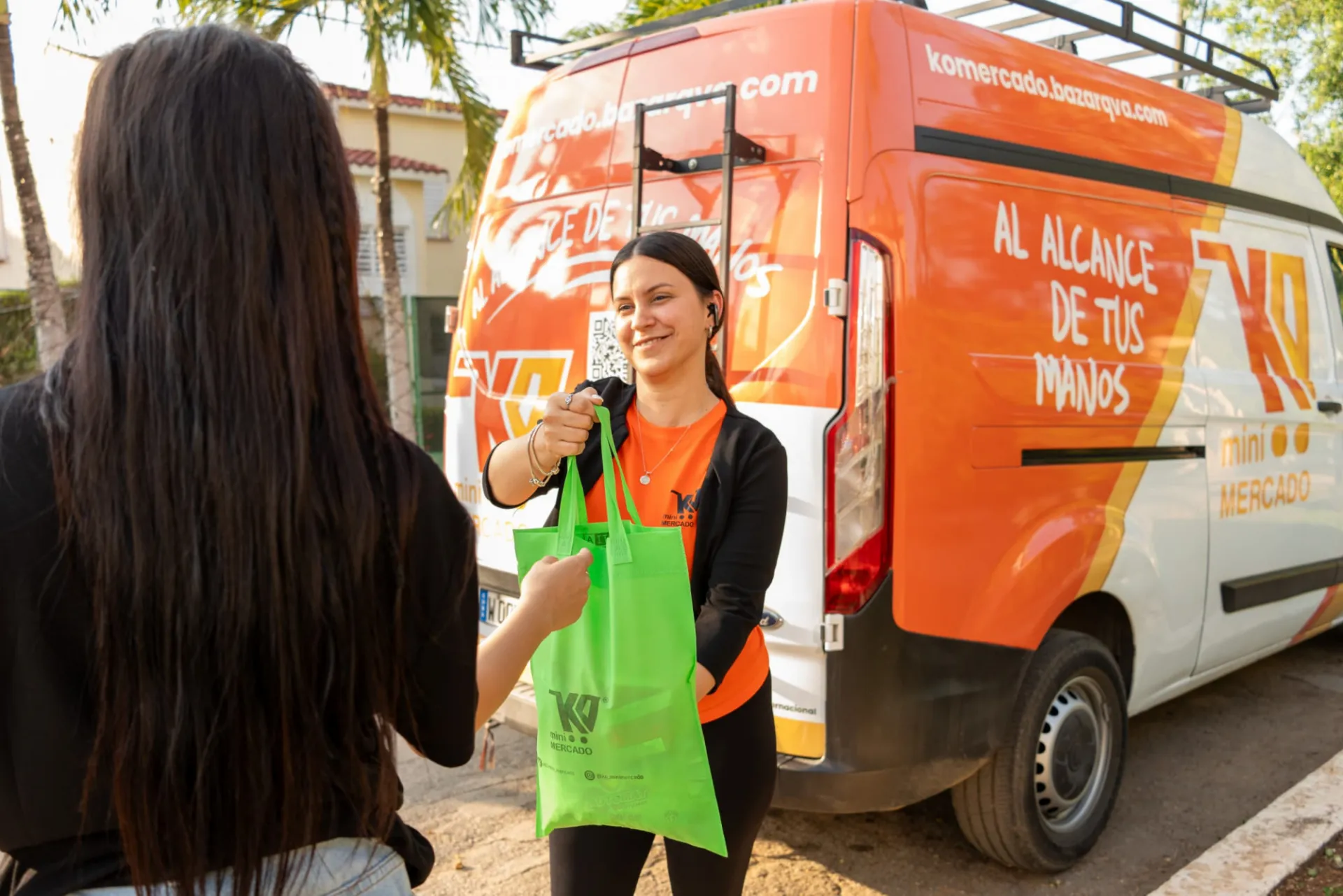 Two women are pictured, one facing away from the camera and the other facing forward. The woman in front is young, wearing an orange T-shirt with a logo, smiling as she hands a green reusable bag to the woman whose back is turned. Behind them, there is a white and orange van with a large sign that reads 'WITHIN YOUR REACH' and 'KO mini MARKET.' The scene takes place outdoors, with trees and buildings in the background, under natural daylight.
