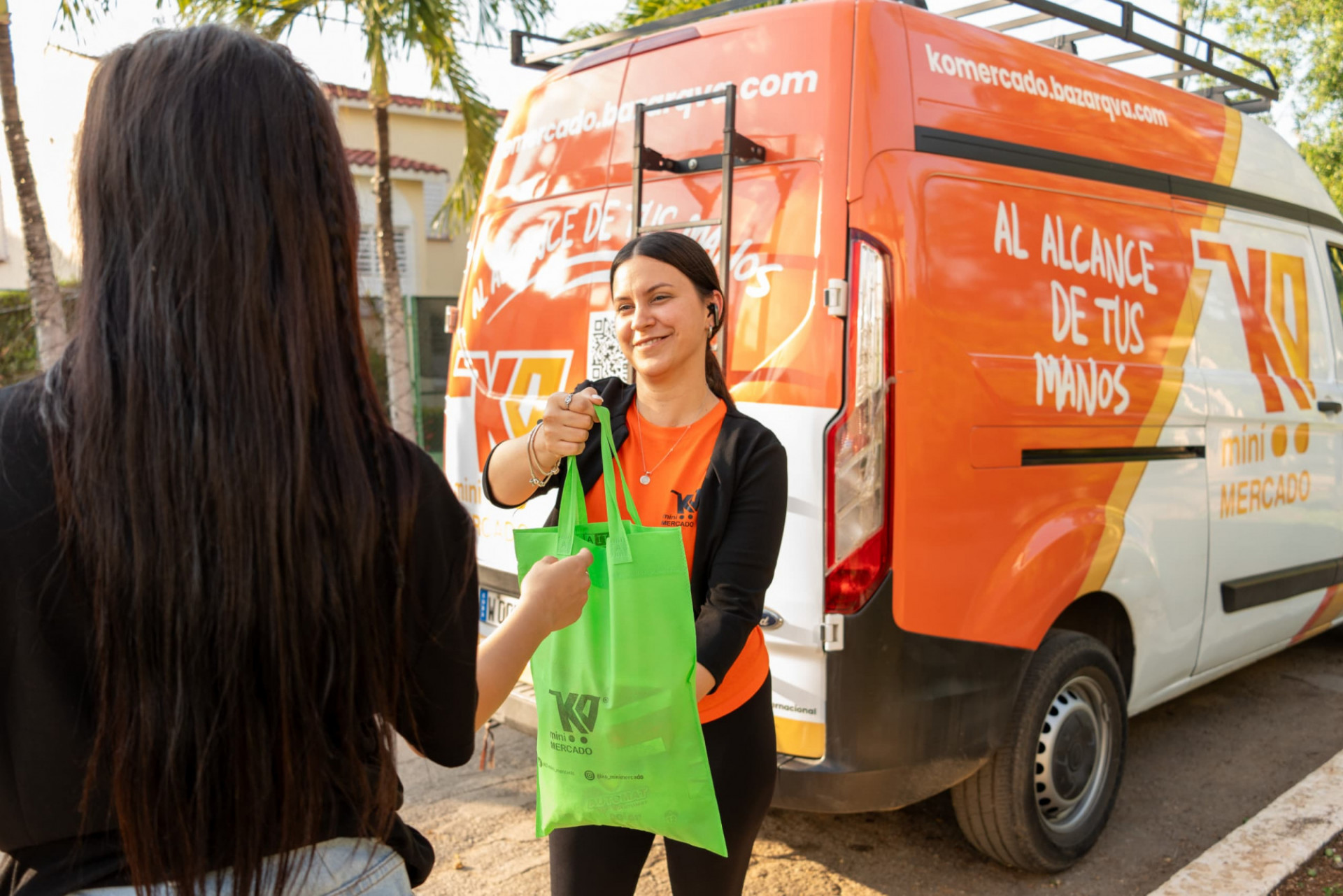 Two women are pictured, one facing away from the camera and the other facing forward. The woman in front is young, wearing an orange T-shirt with a logo, smiling as she hands a green reusable bag to the woman whose back is turned. Behind them, there is a white and orange van with a large sign that reads 'WITHIN YOUR REACH' and 'KO mini MARKET.' The scene takes place outdoors, with trees and buildings in the background, under natural daylight.