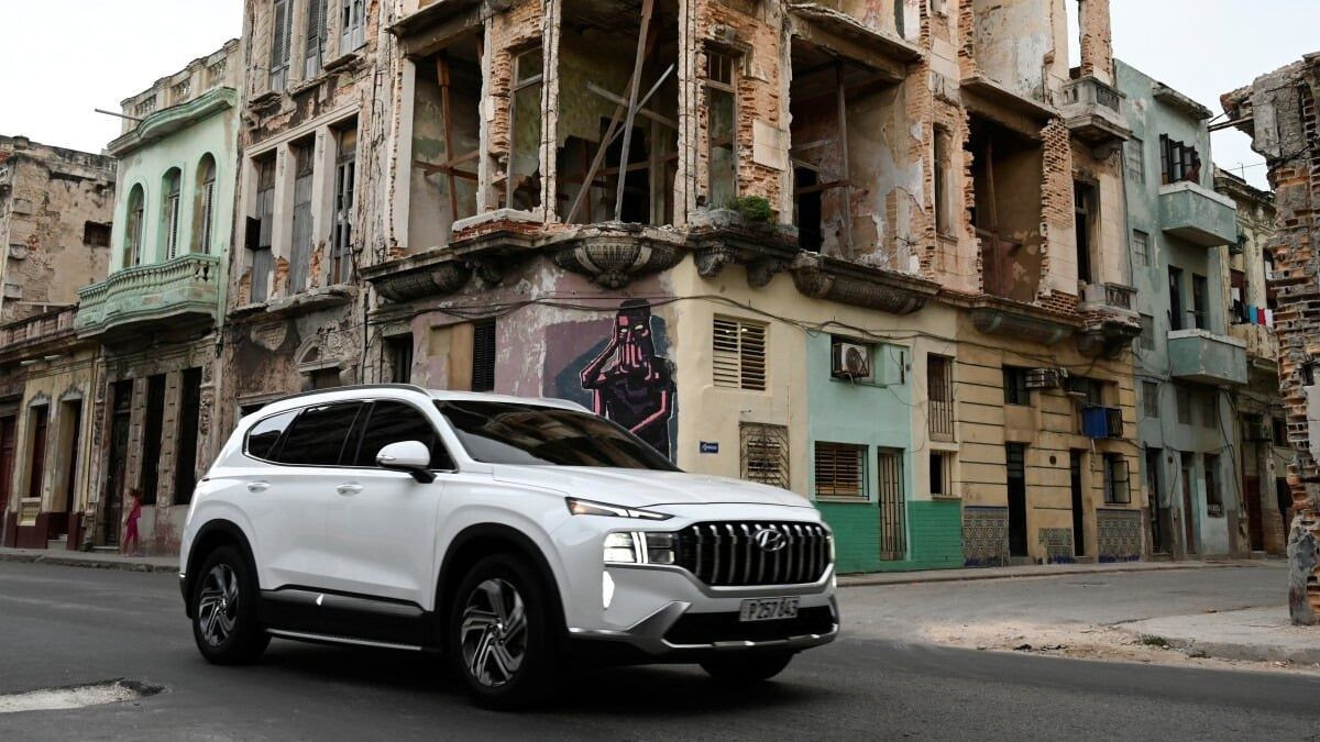 The image shows a modern car driving down a street surrounded by old, dilapidated buildings. The contrast between the new vehicle and the aged facades creates a powerful visual metaphor for the coexistence of modernity and decay in the urban environment.