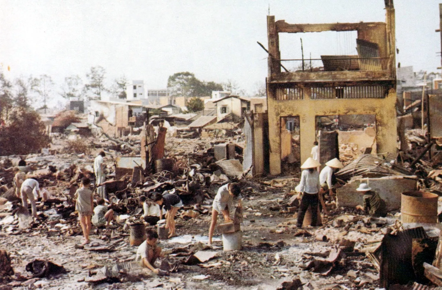 The image depicts a scene of urban destruction. Several people, some wearing traditional conical hats, walk and scavenge among the rubble of collapsed and burned buildings. In the foreground, charred walls, twisted metal sheets, and wooden debris are visible. In the background, a few houses still stand and scattered trees are visible under a clear sky. The overall atmosphere conveys devastation and a struggle to recover what has been lost.