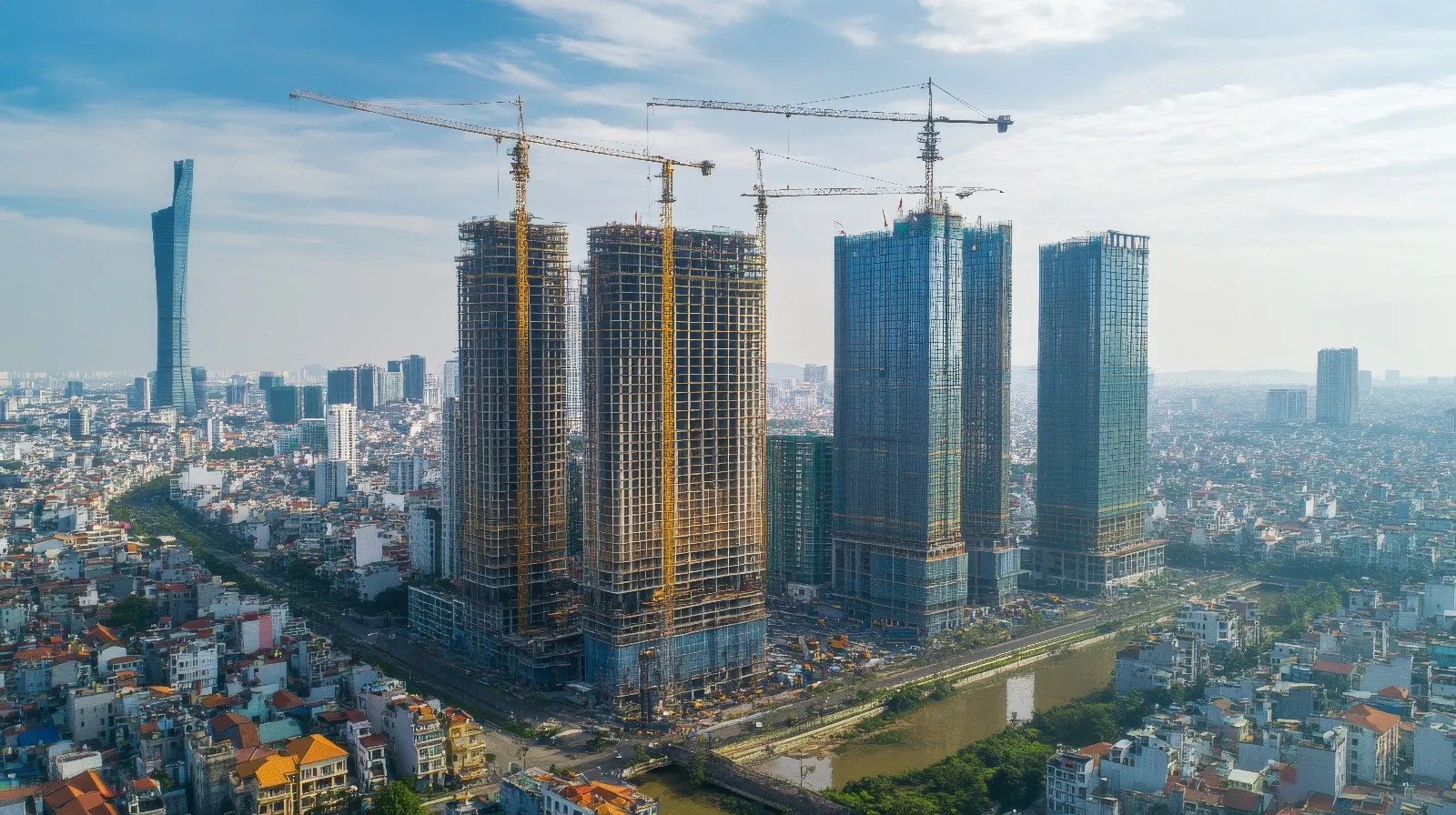 Aerial view of a modern city with several skyscrapers under construction. Five tall towers stand out in the center of the image, surrounded by yellow cranes. The structures are partially covered with scaffolding and blue glass cladding. A dense urban area with low-rise buildings and narrow streets surrounds them. In the background, a skyline filled with more buildings and a clear sky with a few wispy clouds can be seen.