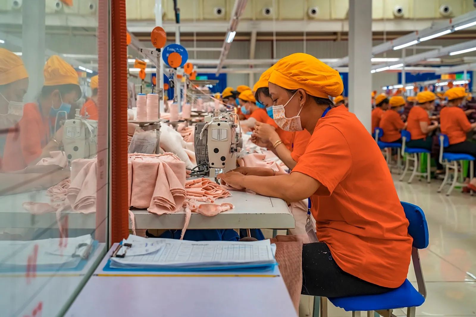 In a spacious, well-lit factory, several people work at a long row of sewing machines. They all wear orange T-shirts, matching caps, and face masks. They are focused on sewing pieces of pink fabric that are piling up on the tables. The atmosphere conveys order, discipline, and industrial productivity.
