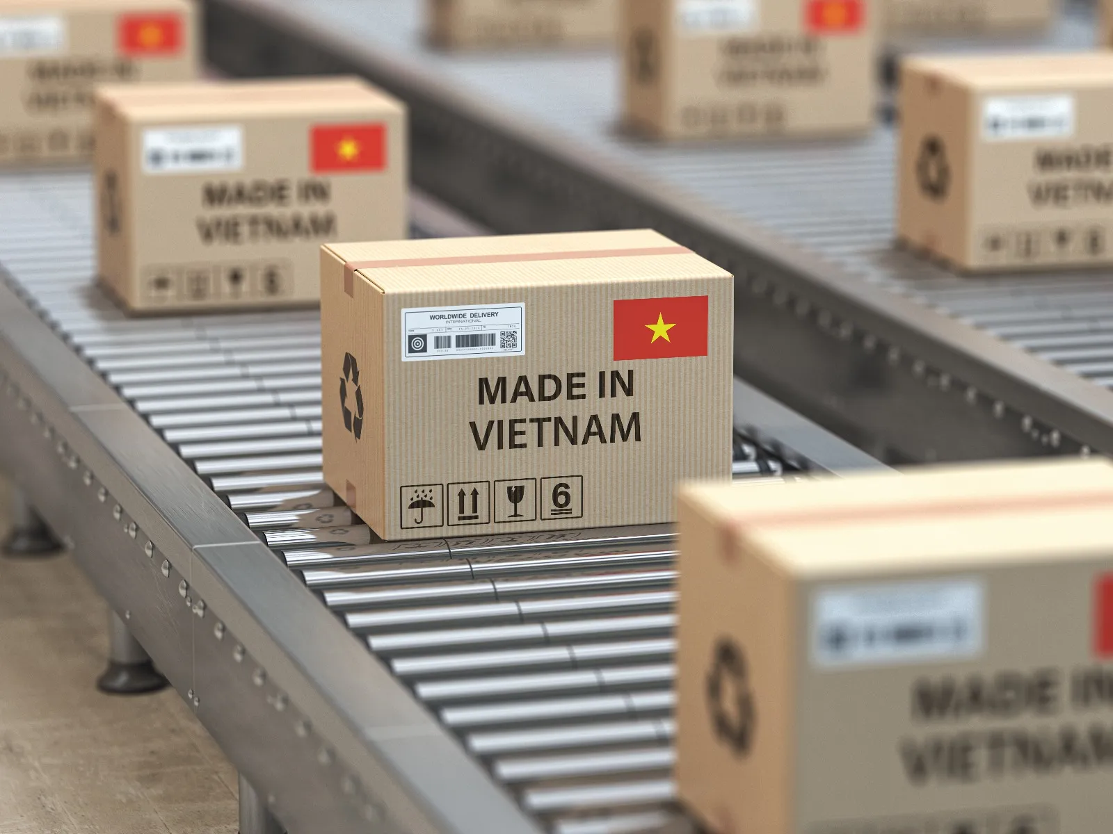 Several cardboard boxes move along a metal conveyor belt on a production line. In the center, a box stands out with the inscription “MADE IN VIETNAM” in black letters and a small Vietnamese flag printed in the upper right corner. The box also displays handling symbols, such as recycling and fragility. In the background, more similar boxes are blurred, suggesting a large-scale packaging or shipping process.